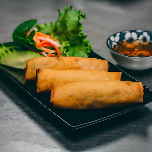 Three golden-brown Vietnamese spring rolls on a black plate with lettuce, carrots, and dipping sauce.