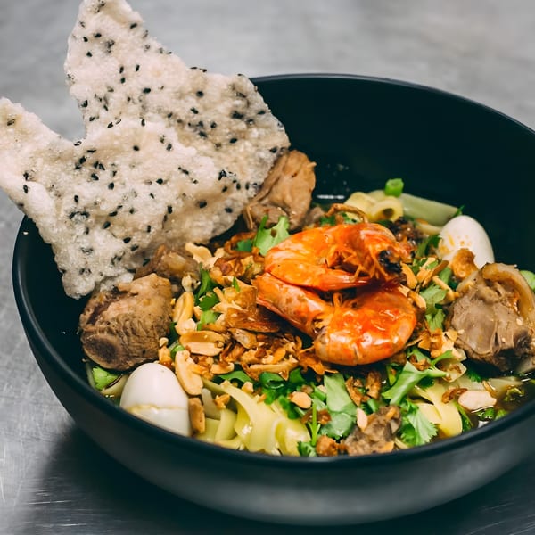 A black bowl of Hên Vietnamese noodles with shrimp, pork, quail eggs, and a sesame rice cracker.