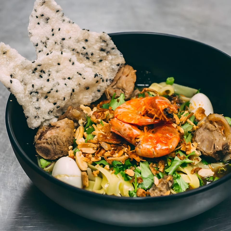A black bowl of Hên Vietnamese noodles with shrimp, pork, quail eggs, and a sesame rice cracker.
