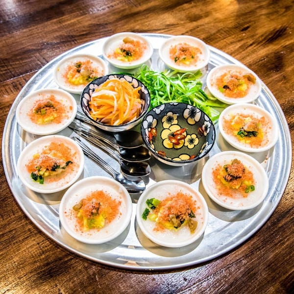 A round silver tray holds small white bowls of Vietnamese Banh Beo, with pickled vegetables and sauce.
