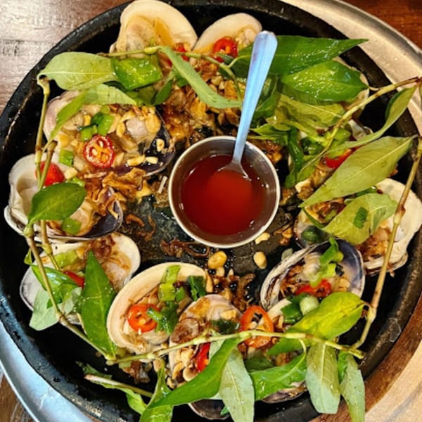 Steamed clams with herbs, peanuts, chili, and a side of red dipping sauce in a black pan.